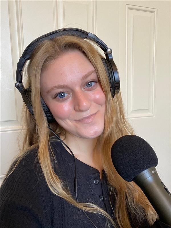 A photo of Kacie Parrow, a student in Saint Leo University's podcast course in the fall of 2020, wearing headphones with a microphone in front of her; she is smiling A photo of Kacie Parrow who was a student in the podcast course at Saint Leo University in the fall of 2020 wearing headphones with a microphone in front of her as she smiles